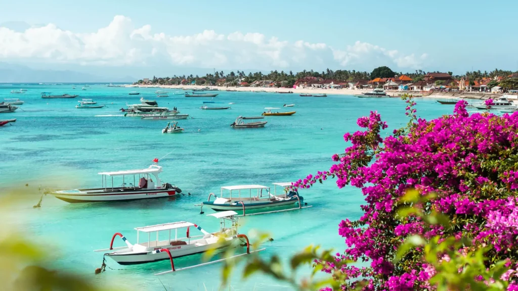 Scenic view of Jungut Batu Beach Nusa Lembongan featuring purple flowers, traditional fishing boats, and fast boats along the shoreline